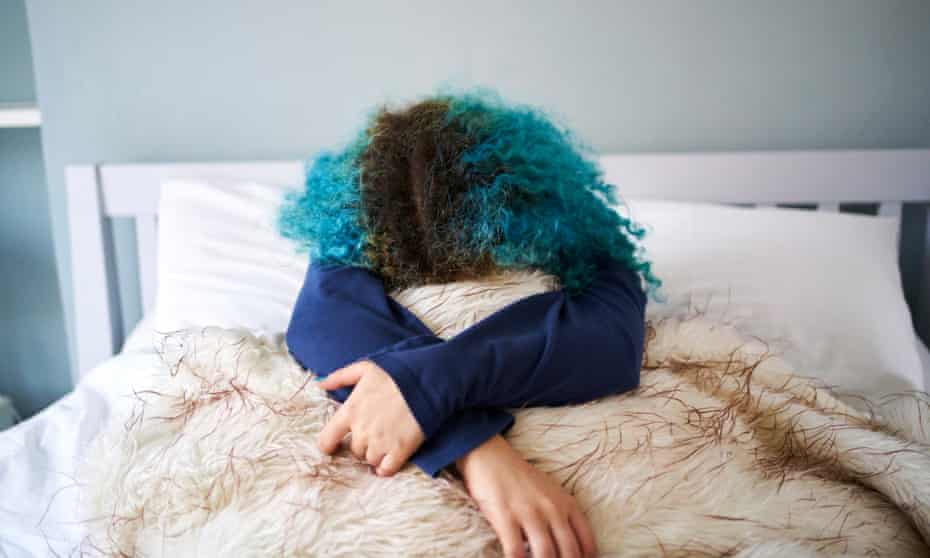 A young woman with bright blue hair and a large streak of regrowth curled up on a bed.
