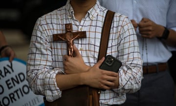 Seen from neck down, white man in collared shirt folds hands on chest holding cellphone in one hand and wooden cross in other.