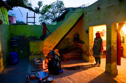A woman kneels by a makeshift stove in a courtyard with metal plates in front of her as another woman stands in the doorway and a child looks on from steps above the yard. It appears to be twilight and the courtyard is in shadow but illuminated from the house.