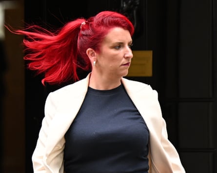 she has bright red hair in a long ponytail, which is billowing behind her; she wears a pale cream jacket and black top, and is seen against a dark background.