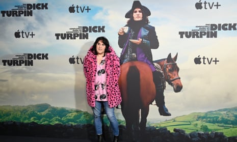 Noel Fielding at the photocall for The Completely Made-Up Adventures Of Dick Turpin, 13 February 2024, London: he is wearing a bright pink fake-fur coat with black spots, a paler pink loose shirt with images of Bugs Bunny, and light blue jeans with black boots, standing in front of a huge poster showing him in costume as Dick Turpin, sitting facing backwards on a horse
