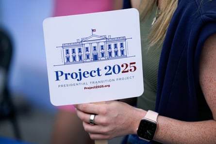 A close-up of a woman’s arm holding up a white sign reading ‘Project 2025’ with an illustration of the White House