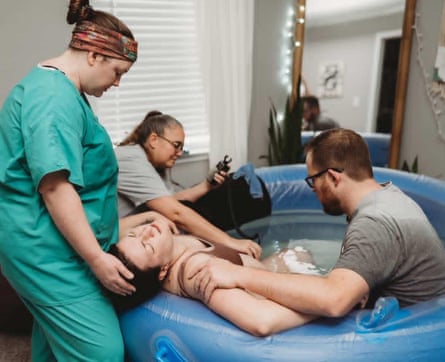 A woman in teal scrubs stands behind a woman sitting in a plastic pool full of water in a living room, holding her head, as a man sits in the pool beside her.