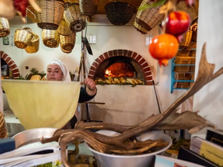In a kitchen, a woman wearing a headscarf lifts a sheet of dough, in the background is a wood fired stove and there are cane baskets hanging from the ceiling