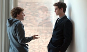 Jesse Eisenberg and Andrew Garfield standing facing each other, talking, in front of a window in The Social Network