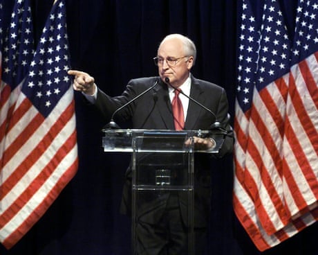a man in suit standing at lectern pointing, several large US flags behind him