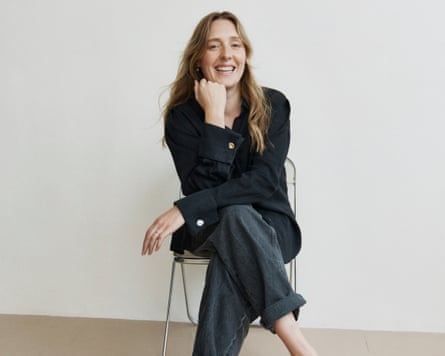 Amy Powney in black shirt and trousers sits on a chair with crossed legs and a hand under her chin.