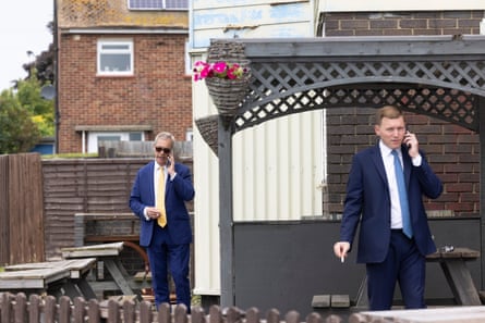 Cottrell and Farage stand apart from each other in a pub beer garden. They are both smoking cigarettes and talking on their mobile phones.