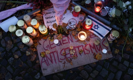 Candles are seen glowing in the twilight along with a small Israeli flag, a bunch of flowers and a sign saying ‘against antisemitism’ are left on cobblestones after a vigil outside a Jewish community centre in Berlin