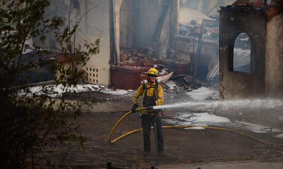 A firefighter in yellow uniform sprays a hose in front of a destroyed, smoking home