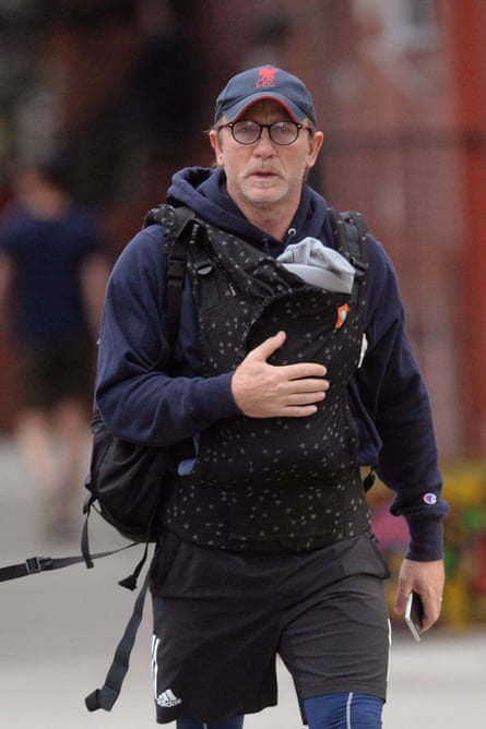 Daniel Craig in casual clothes and using a baby carrier - just visible is the top of a baby's head in a hat