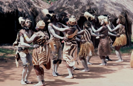 Five couples wearing paint and in traditional skins dance outside huts