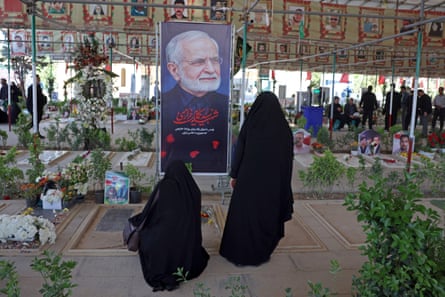 A large portrait of Kharazi is set up in front of a grave in a cemetery with women wearing burqas stood before it