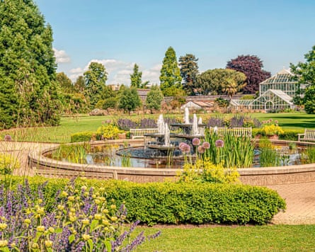 A pond with fountains in a public garden, with trees and greenhouses behind.