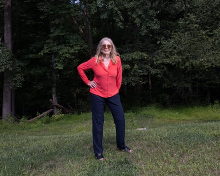 Barbara Res wearing a bright-red shirt and navy trousers, standing with the right hand on her hip outside on grass with trees behind her near her New Jersey home