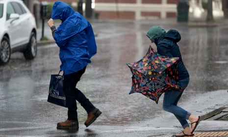 Two people, one in a blue raincoat and the other carrying an umbrella, both covering their heads, cross a very rainy sidewalk.