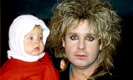 Ozzy Osbourne with backcombed streaked blond hair and heavily made-up eyes, holding baby daughter Kelly in a white bonnet and red coat.
