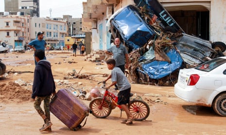 A boy pulling a suitcase and a boy on a bicycle pass wrecked cars piled up against the side of a building