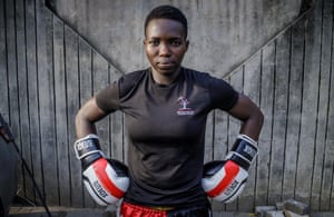 Eniola Taiwo standing in front of tall wood-board fence. She wears a black top and white gloves with red stripe (no headguard).