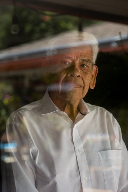 Man with white hair wearing collared white shirt and no tie, appearing to look at camera through a window, with reflections of outdoors across image.