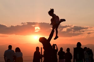 A small crowd are silhouetted against the sunset. A man throws a little girl in the air and catches her
