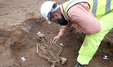 A man in a hard hat and hi-vis clothing bending over a partly unearthed skeleton in the ground