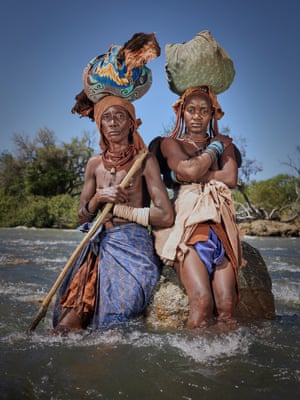 Two women, one older, up to their knees in a fast-flowing river with bundles on their heads