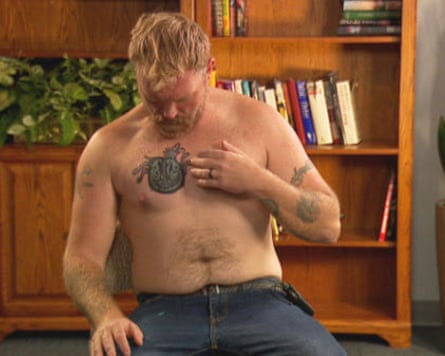 A shirtless man points to a tattoo on his chest