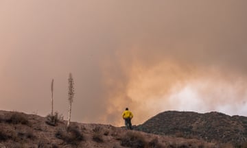 Single man in yellow shirt seen from back on top of hill looking at clouds of smoke.