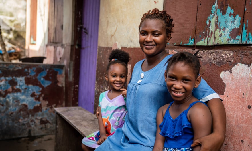 Namissa sitting outside a house with very peeling paint, with her daughters Aminata and Kadijah