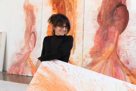 Penny Siopis, wearing glasses and a long-sleeved high-necked shirt, with a fringe and her hair clasped up on her head, grins as she rests her elbow on a canvas in front of three paintings of vertical swirls