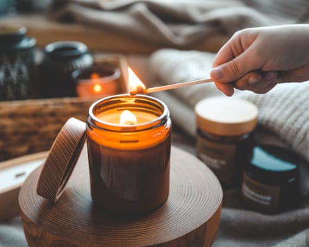 A hand comes into frame to light a candle in glass jar. The background is full of candles and soft furnishings in neutral autumnal hues.
