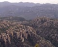 Rock spires rise among forested hills with mountains in background on hazy day