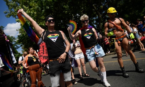 People hold rainbow flags and a fan during a march, while one man is dressed as a scantily clad workman holding a drill