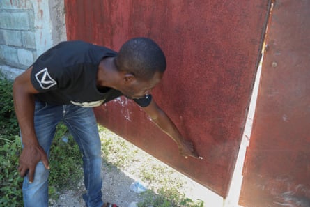 Man in black T-shirt and jeans points to small hole in red metal door.