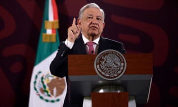 A man wearing a black suit speaks at a podium, with the Mexican flag hanging behind him