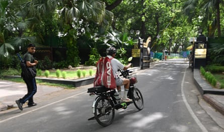 An Indian man on a motorbike and another on foot, both carrying a bag with squash racquets, approach a gate in a tree-lined residential area