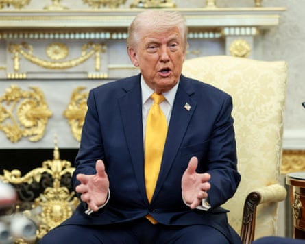 he holds his hands out as he speaks. He wears a dark blue suit with a yellow tie and a gold-embellished fireplace and gold ornaments are in the background.