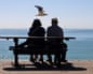 An elderly couple sit on a bench looking out across a beach to the sea. Above them hovers a seagull.