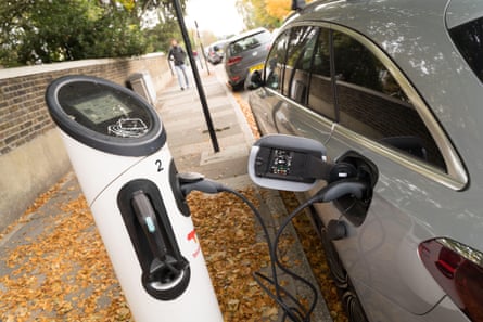An electric vehicle is plugged into a roadside charging point on a pavement covered with autumn foliage