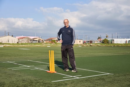 An older white man stands on a cricket pitch with low-rise buildings in the background