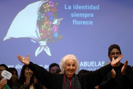 An older woman with white hair raises both arms up in a gesture of celebration