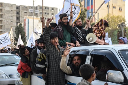 Afghanistan men and boyd cheer, standing on the back of a 4x4 vehicle in a city.