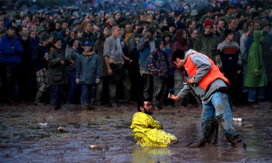 A festival-goer offers a cigarette to a man in a high vis suit who sits in a puddle of mud as a crowd watches the action on stage.