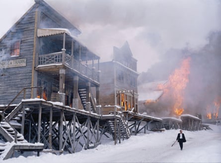A western scene showing a fire raging through a settlement town, while a small figure in black and white walks through the snow