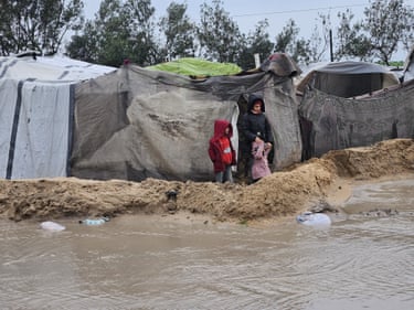 Two people are visible outside tents after heavy rains flood thousands of tents sheltering displaced civilians in Gaza