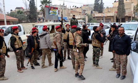 A group of fighters stands in a street; they wear matching bright red berets and fatigues, and carry guns, but some also wear civilian jackets and trainers; several look very young