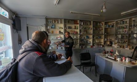 A pharmacists points to products on poorly-stocked shelves while customers wait at a counter.