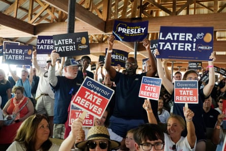 Smiling crowd wave blue and white signs, and red and white signs, all beneath a large indoor-outdoor wooden roof.