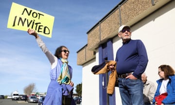 A woman, left, holds a yellow sign reading 'Not Invited' while a man beside her looks off to the side.
